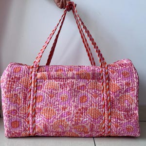 May include: A rectangular quilted duffel bag featuring a vibrant floral pattern in shades of pink, orange, and white. The bag includes two long, sturdy handles with a red and white striped design, providing a secure grip for carrying the item.