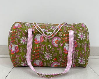 May include: A quilted duffel bag featuring an olive green base with a repeating floral pattern in shades of pink, white, and orange. The bag includes two long, striped pink and white fabric handles and a matching pink and white piped trim.