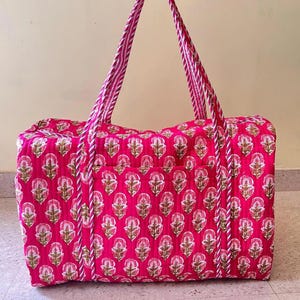 May include: A quilted duffel bag in a vibrant hot pink color featuring a repeating floral block print pattern in white and muted green. The bag includes two long, sturdy carrying handles with a white and pink striped design.