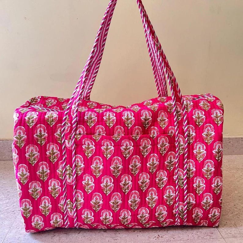 May include: A quilted duffel bag in a vibrant hot pink color featuring a repeating floral block print pattern in white and muted green. The bag includes two long, sturdy carrying handles with a white and pink striped design.