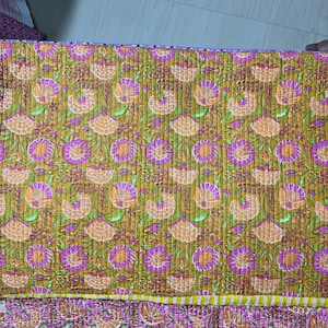 Puede incluir: Una tela acolchada y plegada con un estampado floral. El diseño presenta flores repetidas en tonos naranja y morado sobre un fondo amarillo mostaza. Hojas y tallos verdes se intercalan en todo el diseño. La tela se utiliza probablemente para ropa de cama o decoración del hogar.
