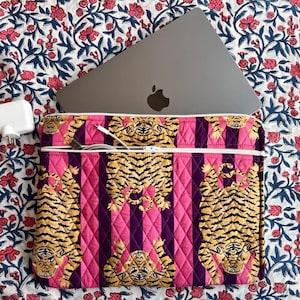 May include: A quilted laptop sleeve featuring a repeating pattern of tigers over vertical pink and dark purple stripes. The sleeve has a white zippered pocket on the front containing a white charging cable, with a silver laptop partially inserted.