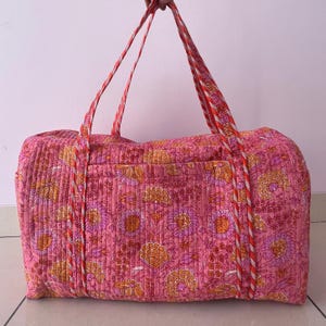 Peut inclure: Grand sac de voyage matelassé rose vif, orné d'un motif floral aux tons orange et violet. Le sac est doté de poignées tressées robustes avec des accents rouges et blancs, offrant un design élégant et pratique.
