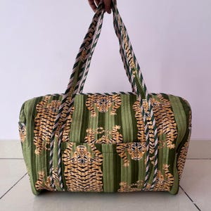 May include: A quilted duffel bag featuring a repeating pattern of tigers on a green background with vertical stripes. The bag has two long handles with a striped pattern and an exterior pocket on the front. The fabric has a stitched texture.