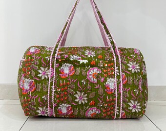 May include: A quilted duffel bag featuring an olive green base with a vibrant floral pattern in shades of pink, orange, and white. The bag includes two long, striped pink and white fabric handles and a rectangular shape with visible stitching.