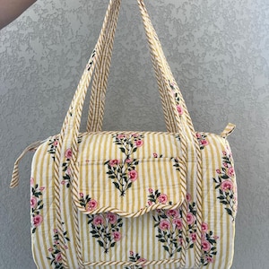 May include: A floral print tote bag with a yellow and white striped pattern. The bag features pink flowers and green leaves. The bag has two handles and a small pocket on the front. The bag is approximately 12 inches wide and 8 inches tall.
