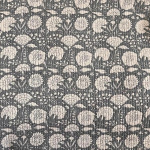 May include: A quilted fabric with a repeating floral pattern. The design features large, stylized white flowers and foliage against a dark gray background. The fabric has a textured appearance due to the quilting stitches, creating a tactile and visually appealing surface.