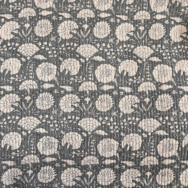 May include: A quilted fabric with a repeating floral pattern. The design features large, stylized white flowers and foliage against a dark gray background. The fabric has a textured appearance due to the quilting stitches, creating a tactile and visually appealing surface.