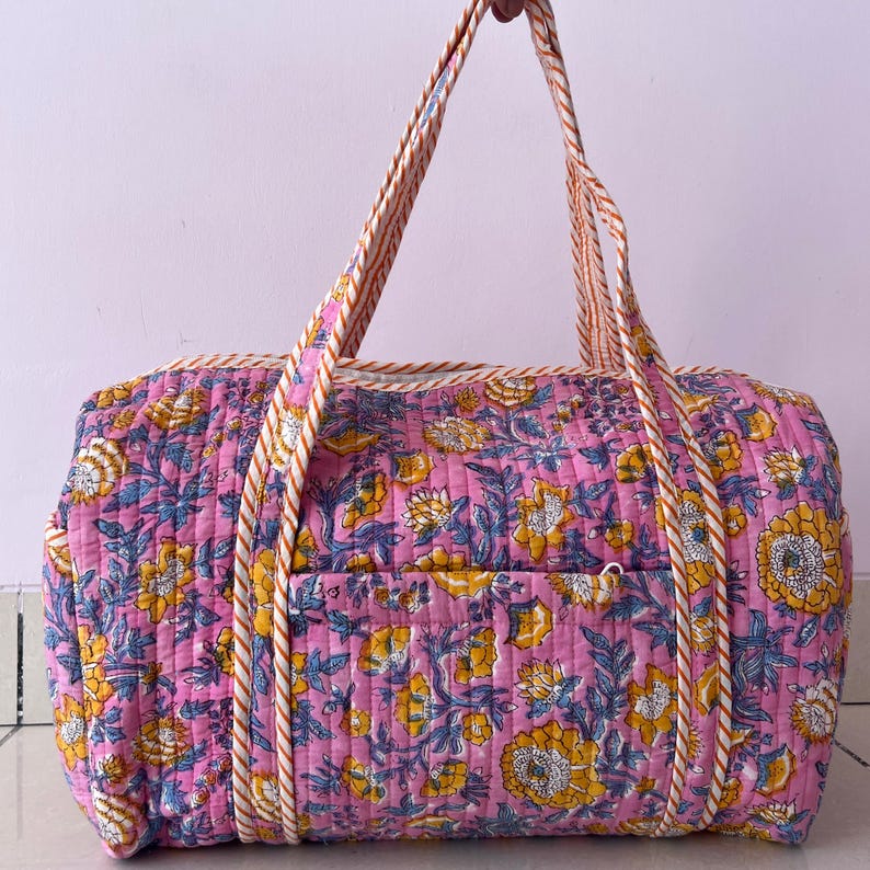May include: A quilted duffel bag featuring a vibrant pink base with a repeating floral pattern in shades of yellow, white, and blue. The bag includes two long, striped fabric handles and a rectangular front pocket with a horizontal quilted texture.