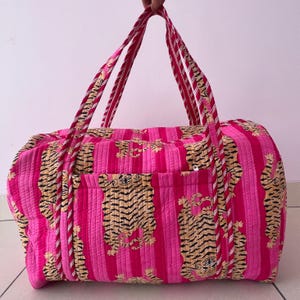 May include: A quilted duffel bag featuring a vibrant hot pink and light pink vertical striped pattern with a repeating tiger print. The bag includes two long, striped fabric handles and a horizontal exterior pocket with a visible white zipper.
