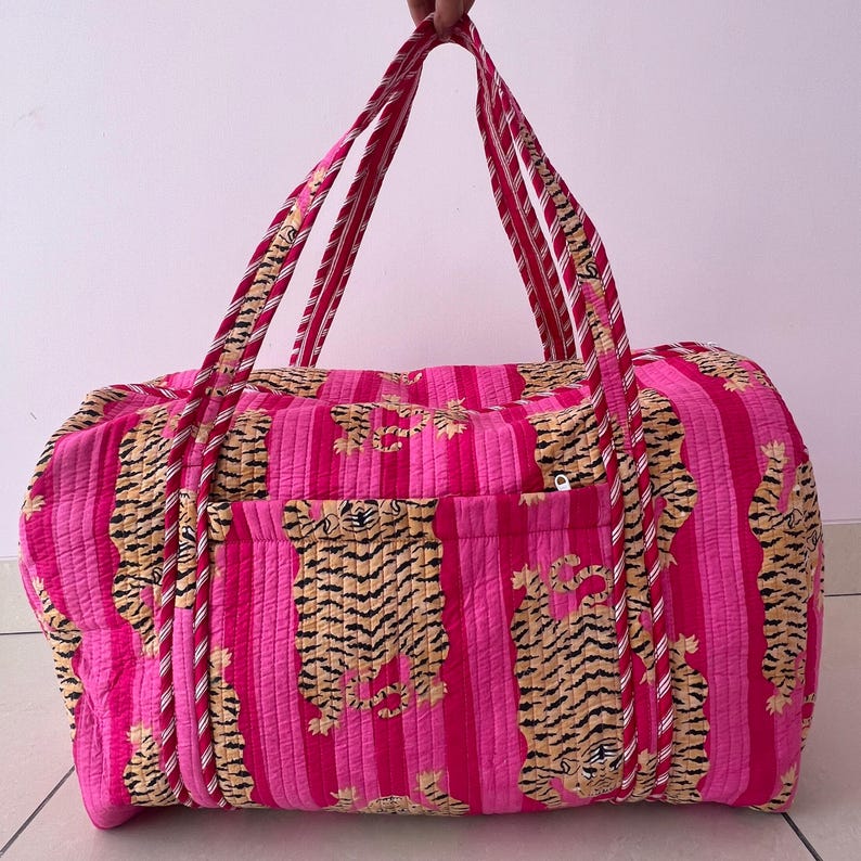 May include: A quilted duffel bag featuring a vibrant hot pink and light pink vertical striped pattern with a repeating tiger print. The bag includes two long, striped fabric handles and a horizontal exterior pocket with a visible white zipper.
