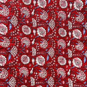 May include: Close-up of a red fabric with a floral pattern. The design features white and pink flowers, blue leaves, and dark stems. The fabric has a quilted texture, creating a visually rich and detailed surface.