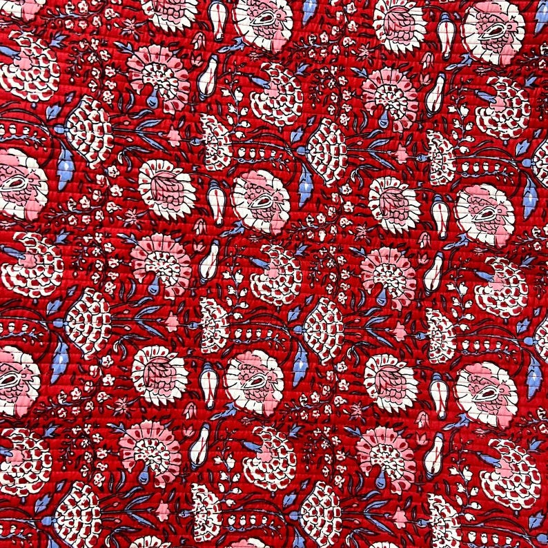 May include: Close-up of a red fabric with a floral pattern. The design features white and pink flowers, blue leaves, and dark stems. The fabric has a quilted texture, creating a visually rich and detailed surface.