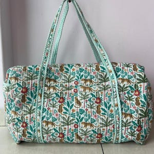 May include: A quilted duffel bag featuring a repeating pattern of leopards, palm trees, and red flowers on a white background. The bag includes two long, light green and white striped fabric handles and a rectangular, soft-sided structure.