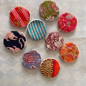 Puede incluir: Nueve monederos redondos de tela con varios patrones florales y geométricos coloridos. Los monederos tienen cremalleras blancas y están acolchados.