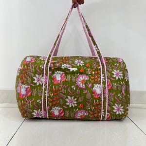 May include: A quilted duffel bag featuring an olive green base with a vibrant floral pattern in shades of pink, orange, and white. The bag includes two long, striped pink and white fabric handles and a rectangular shape with visible stitching.