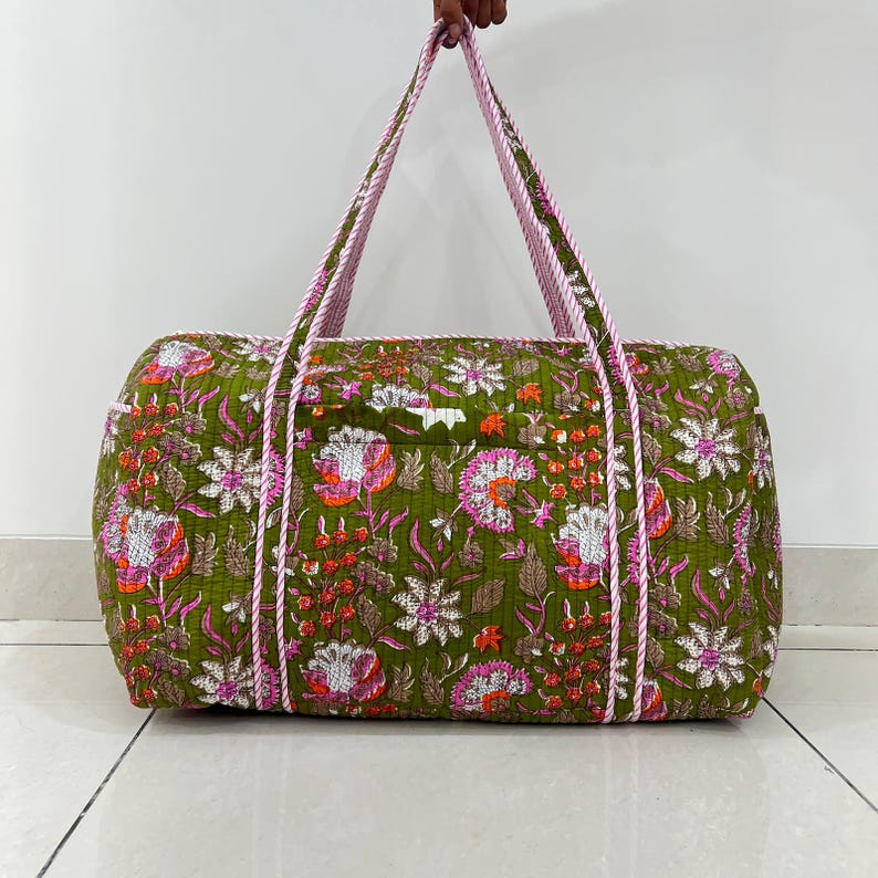 May include: A quilted duffel bag featuring an olive green base with a vibrant floral pattern in shades of pink, orange, and white. The bag includes two long, striped pink and white fabric handles and a rectangular shape with visible stitching.