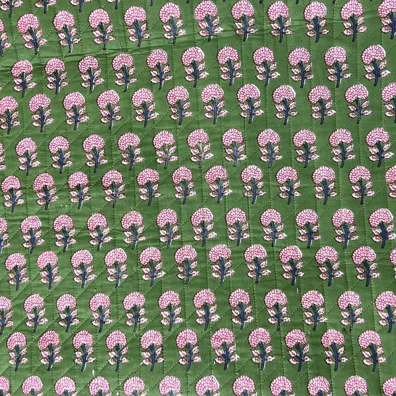 May include: Green fabric with a repeating pattern of pink floral designs. The flowers have a detailed, textured appearance, and the fabric has a quilted effect. The overall design is symmetrical and visually appealing.