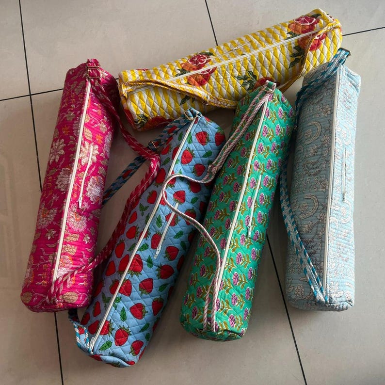 May include: A collection of colorful yoga mat bags. The bags feature various patterns, including floral, strawberry, and geometric designs. Each bag has a zipper closure and a rope handle. The bags are in shades of pink, blue, green, and yellow.