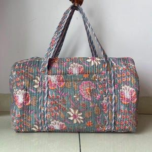 May include: A rectangular quilted duffel bag featuring a muted sage green base with a repeating floral pattern in shades of pink, white, and brown. The bag includes two long shoulder straps and a horizontal exterior pocket across the front panel.