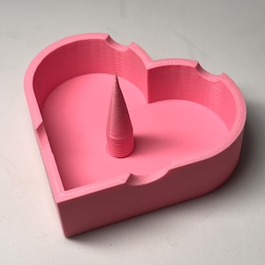 May include: A pink heart-shaped dish with a small pink cone in the center. The dish is made of plastic and has a smooth surface.