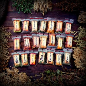 May include: A collection of small, clear plastic bags filled with dried herbs and flowers. Each bag has a label with the name of the herb or flower written on it. The labels are in a vintage style font and are printed on white paper. The bags are arranged in a grid pattern on a wooden surface.