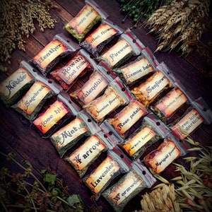 May include: A collection of small, clear plastic bags filled with dried herbs and flowers. Each bag has a handwritten label with the name of the herb or flower, such as "Mottle", "Heather", "Rowan dried fruits", "Fireweed", "Calendula", "Jasmine flower", "Rose petals", "Sweet basil", "Lemon balm", "Sanfiowe", "Dandelion flower", "Wormwoo", "Marigold flower", "ree lobe gartick", "Tamin sandy", "lac flowe", "Sagebrush", "Rose hip", "Mint", "arrow", "Lavender".