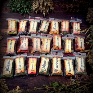 May include: A collection of 18 small, clear plastic bags filled with dried herbs and flowers. Each bag has a label with the name of the herb or flower written on it. The labels are in a vintage style with brown and white lettering. The bags are arranged in three rows of six on a wooden surface.