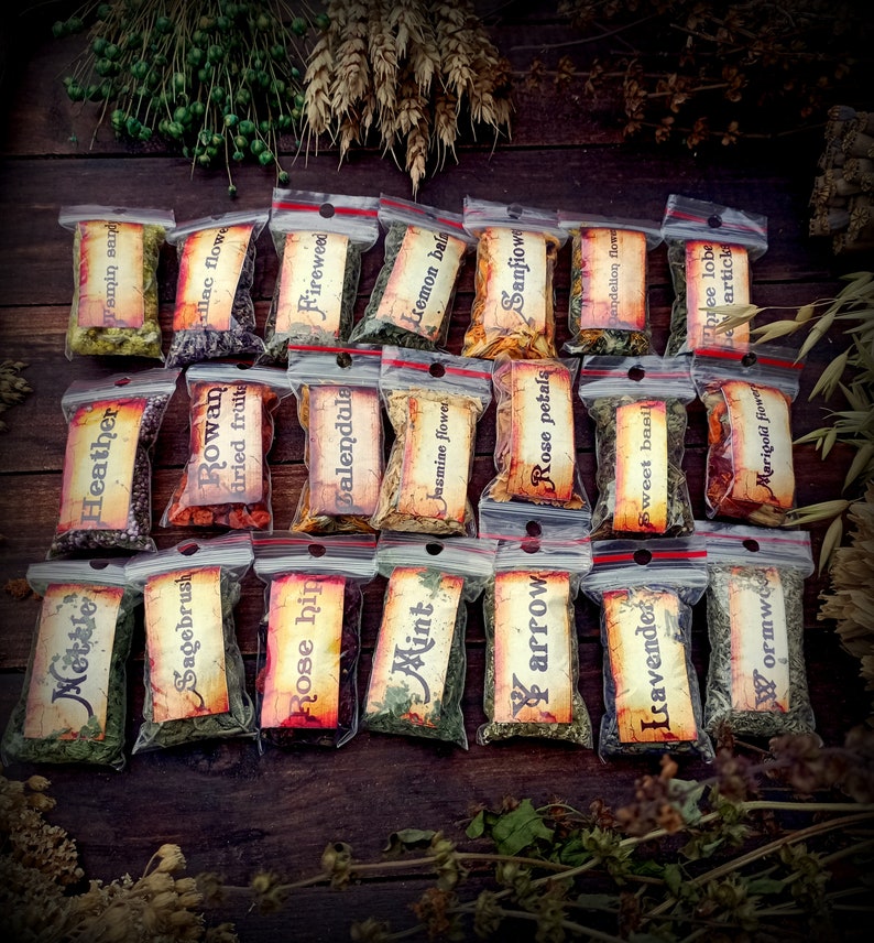 May include: A collection of 18 small, clear plastic bags filled with dried herbs and flowers. Each bag has a label with the name of the herb or flower written on it. The labels are in a vintage style with brown and white lettering. The bags are arranged in three rows of six on a wooden surface.