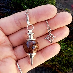 Könnte beinhalten: Ein Pendel mit silberner Kette und Details. Das Pendel hat einen braunen, gesprenkelten, kugelförmigen Stein und eine silberne konische Spitze. Ein silberner Anhänger mit einem vierzackigen Design hängt an einer separaten Kette.