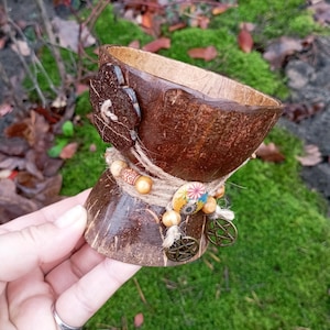 Puede incluir: Un cuenco artesanal de cáscara de coco con un diseño decorativo. El cuenco es marrón con una textura natural, adornado con hilo, cuentas de madera y abalorios metálicos. Una pieza similar a un botón asegura la parte superior. El cuenco se sostiene en una mano.