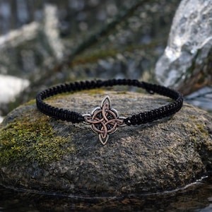 May include: A black macrame choker necklace with a silver and copper Celtic knot pendant. The necklace is displayed on a moss-covered rock, with a blurred background of water and snow.