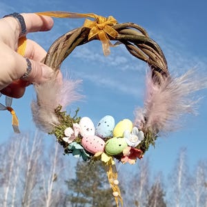 May include: A small, twig wreath decorated with pastel colored Easter eggs, pink feathers, and yellow ribbon.