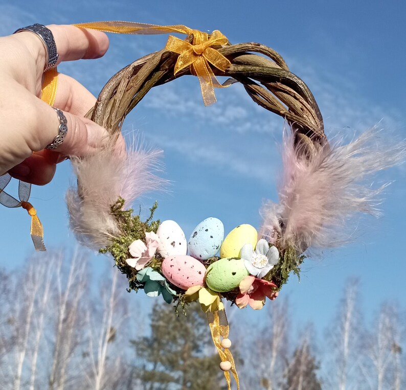 May include: A small, twig wreath decorated with pastel colored Easter eggs, pink feathers, and yellow ribbon.