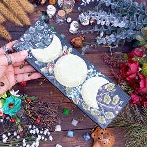 May include: A dark blue wooden key holder featuring three moon phases with floral and botanical accents. The key holder has brass hooks and is decorated with white, green, and red details. The key holder is approximately 12 inches long.