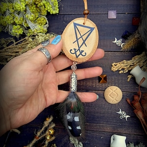 Op de afbeelding: Een houten hanger met een zwart sigil erop geschilderd. De hanger is bevestigd aan een bruin leren koord en een zilveren verenbedel. De hanger is omringd door verschillende kruiden en andere magische voorwerpen.