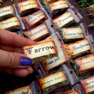 May include: A collection of small, clear plastic bags filled with dried herbs and flowers. Each bag has a handwritten label with the name of the herb or flower, such as "Yarrow", "Calendula", "Fireweed", and "Rose petals".
