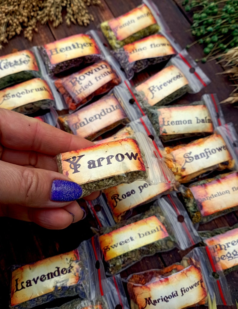 May include: A collection of small, clear plastic bags filled with dried herbs and flowers. Each bag has a handwritten label with the name of the herb or flower, such as "Yarrow", "Calendula", "Fireweed", and "Rose petals".
