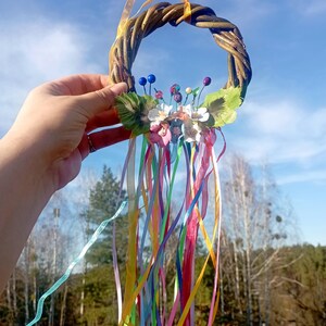 May include: A woven willow wreath with colorful ribbons and fabric flowers. The wreath is decorated with blue, pink, and white flowers and green leaves. The ribbons are a variety of colors, including pink, yellow, blue, green, and purple.