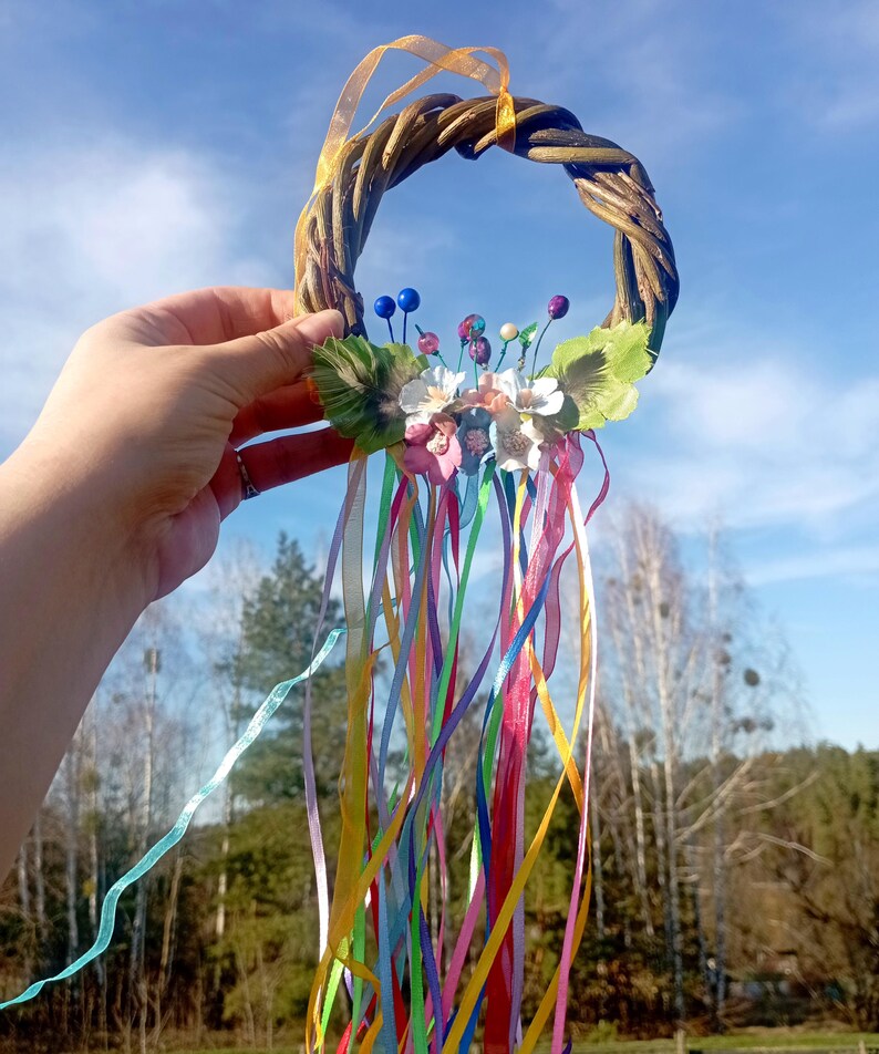 May include: A woven willow wreath with colorful ribbons and fabric flowers. The wreath is decorated with blue, pink, and white flowers and green leaves. The ribbons are a variety of colors, including pink, yellow, blue, green, and purple.