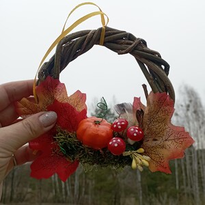 May include: A small autumn wreath made of twigs with red and orange fall leaves, a small orange pumpkin, and red and white mushrooms.