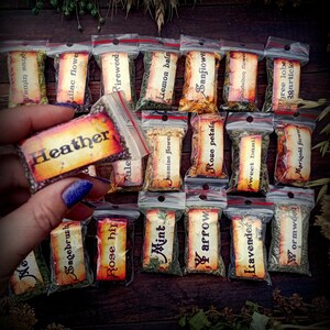 May include: A collection of small, clear plastic bags filled with dried herbs and flowers. Each bag has a label with the name of the herb or flower written on it. The labels are in a vintage style with a brown background and white lettering. The herbs and flowers include: Heather, Sagebrush, Rose hip, Mint, Lemon balm, Jasmine flower, Sanfiowe, arrow, Rose petals, Dandelion flower, Isavender, Sweet basil, Wormwood, Phree lobe, 3garticks, Marigold flower.