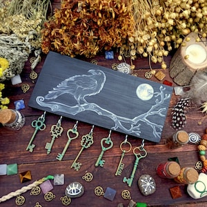 May include: A black wooden key holder featuring a white raven and branch design. The holder has six hooks for hanging keys. The background includes various herbs, crystals, and other witchy decor.