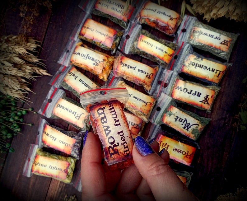 May include: A variety of small, clear plastic bags filled with dried herbs and flowers. Each bag has a label with the name of the herb or flower written in a rustic font. The bag in the center of the image is labeled "Rowan dried fruits".