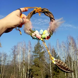 May include: A handmade Easter wreath made of twigs, decorated with pastel colored eggs, a pink feather, and a yellow ribbon. The wreath is hanging from a string of beads and feathers.