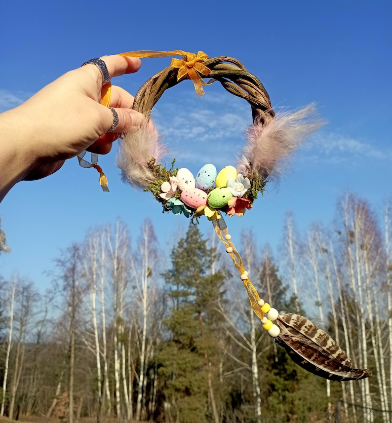 May include: A handmade Easter wreath made of twigs, decorated with pastel colored eggs, a pink feather, and a yellow ribbon. The wreath is hanging from a string of beads and feathers.