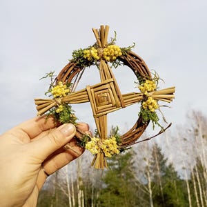 May include: A woven straw wreath with a cross design, decorated with dried yellow flowers and green moss.