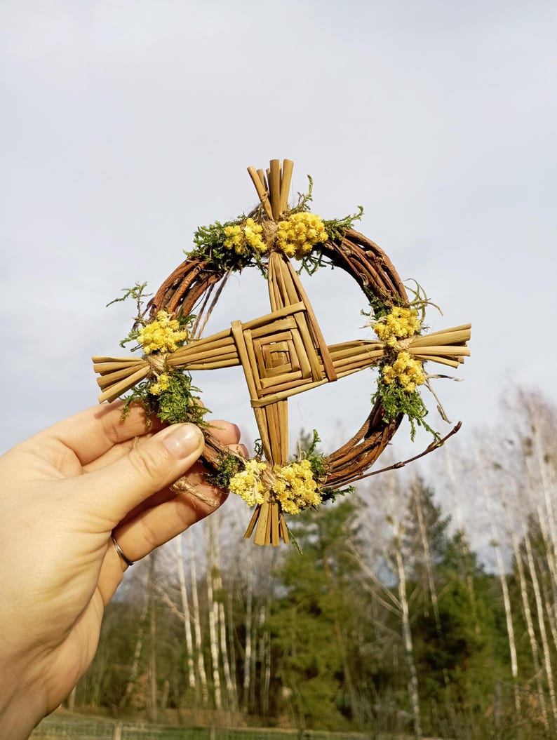 May include: A woven straw wreath with a cross design, decorated with dried yellow flowers and green moss.