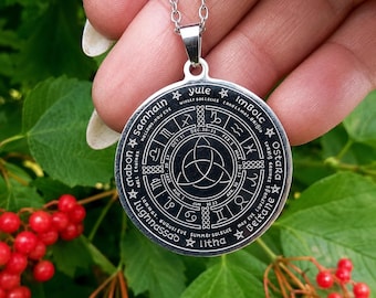 May include: A silver-toned circular pendant on a matching chain featuring a black engraved wheel of the year. The design includes the names of eight sabbats, zodiac symbols, dates, and a central triquetra knot symbol in a detailed circular layout.