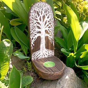 May include: A wooden candle holder with a tree design. The dark brown holder features a white tree illustration and a circular base holding a green candle. The background includes green foliage and a gray rock.